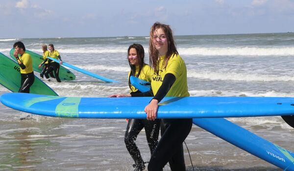 Twee kinderen lopen uit de zee met een surfplank tijdens een kindervakantie. De criteria van het keurmerk van kindervakanties vind je hier.