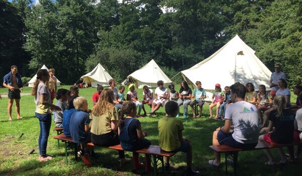 Kinderen en volwassen zitten in een kring bij elkaar tijdens een kindervakantie. Kindervakantieorganisaties kunnen een keurmerk aanvragen via Jantje Beton.