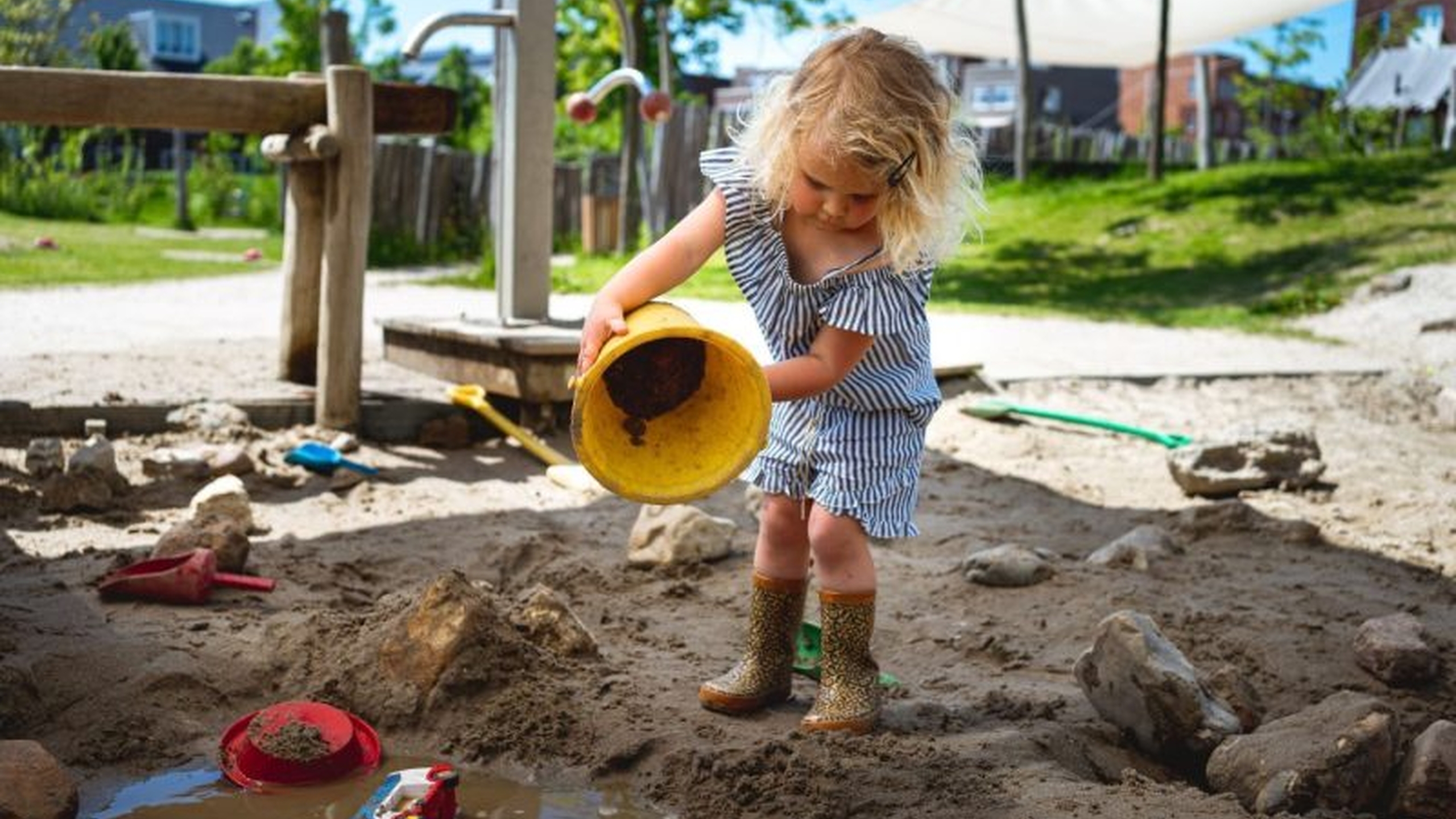 Een kindje gooit zand uit een emmer. Het is belangrijk om erkenning en financiering van gemeenten te krijgen voor speeltuinen - Jantje Beton